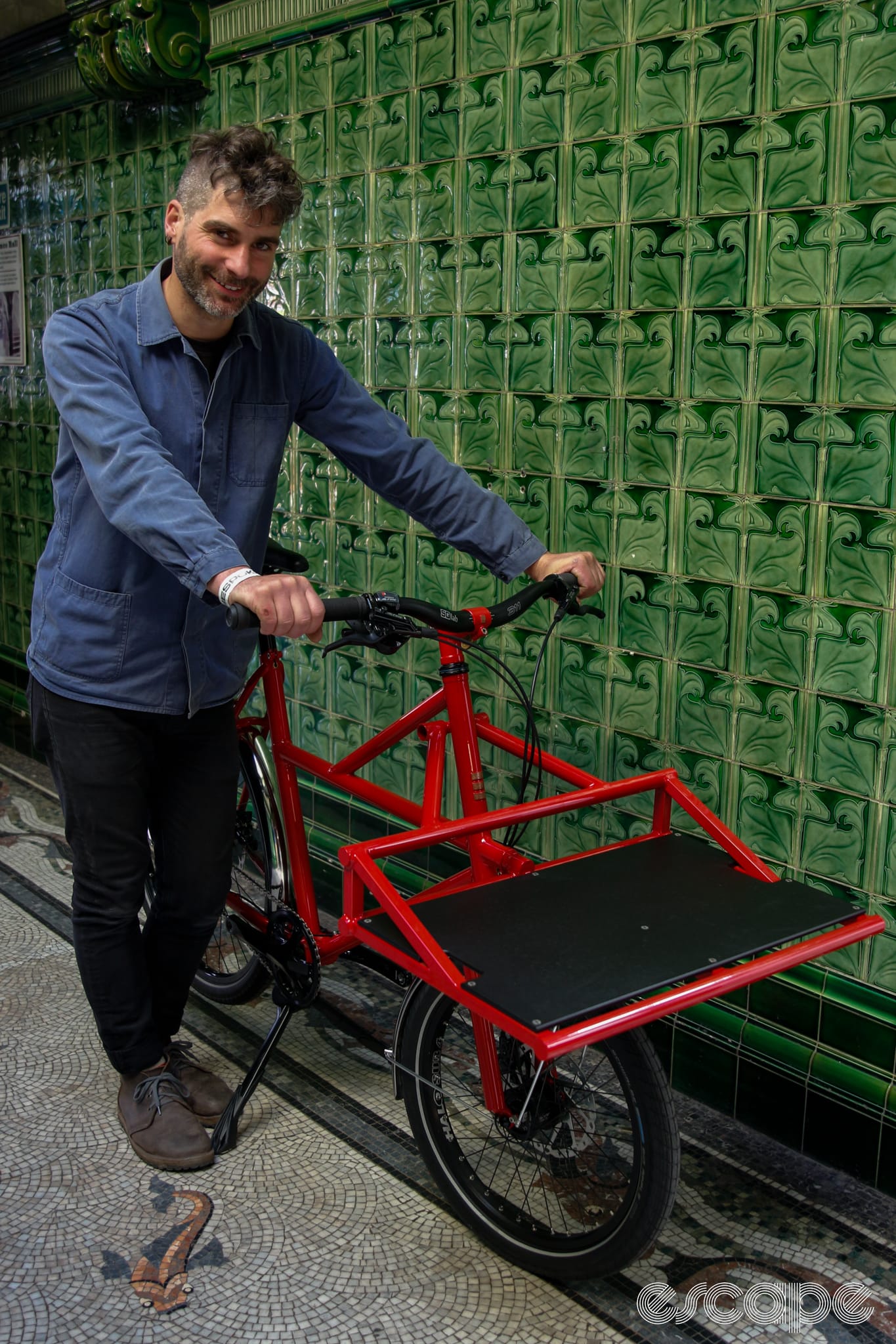 Pi Manson of Clandestine with his red small cargo bike, against green tiled wall.
