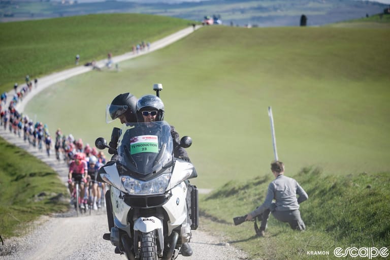 A television camera moto leads the pack at the 2023 Strade Bianche. The cameraman is peering through his eyepiece with the ca